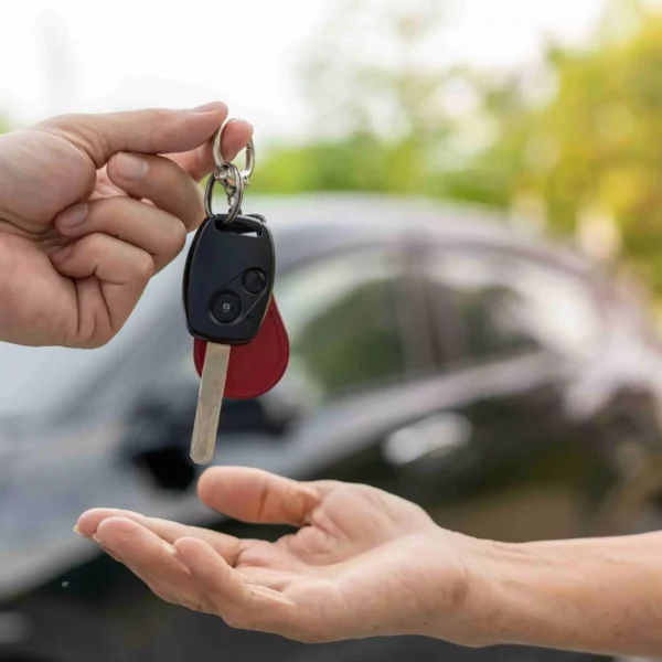 A hand handing over car keys to another person with a car in the background.